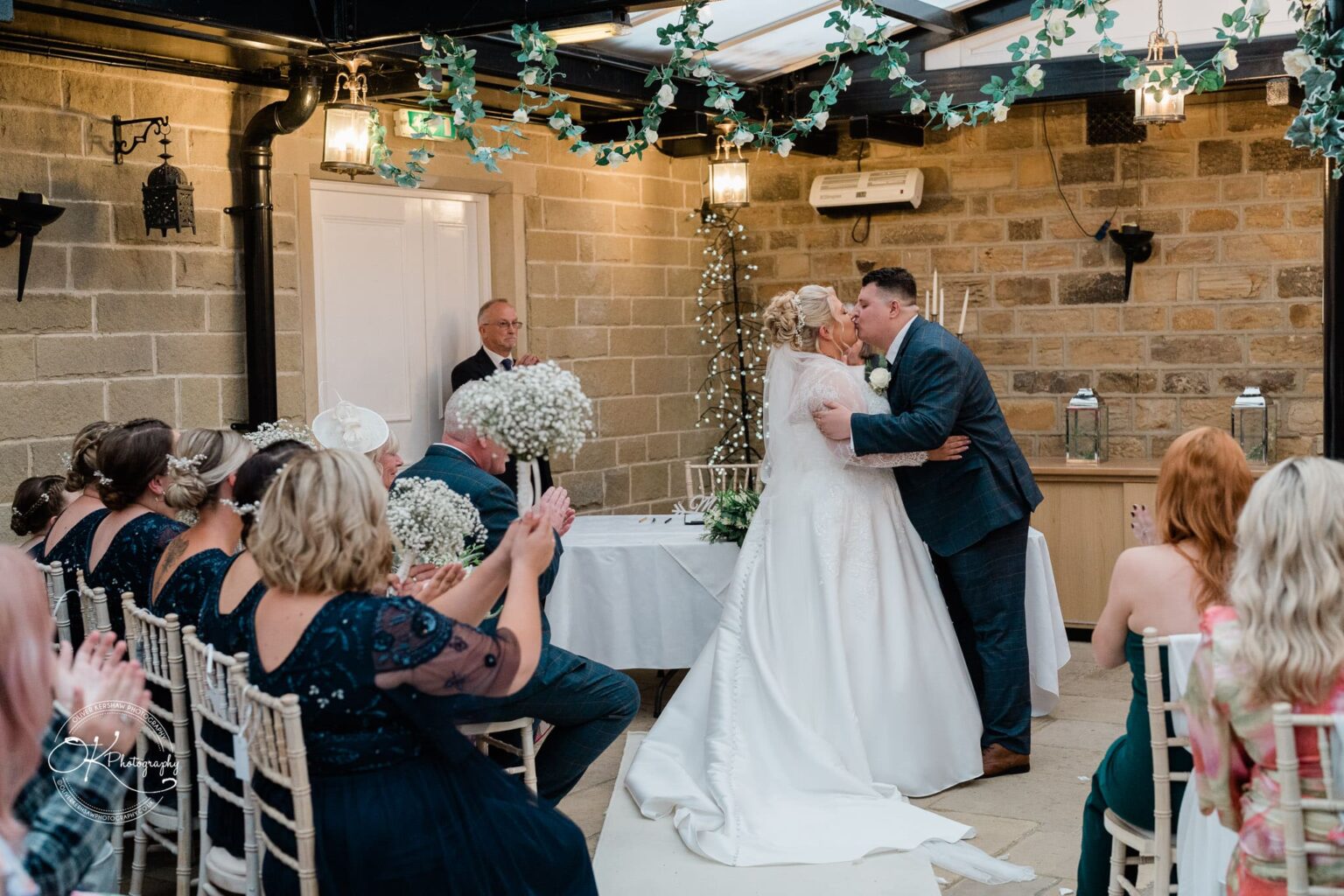 Santos Higham Farm Wedding Photography A bride and groom share a kiss during their wedding ceremony at Santos Higham Farm, surrounded by guests who are clapping and holding bouquets of baby's breath flowers.