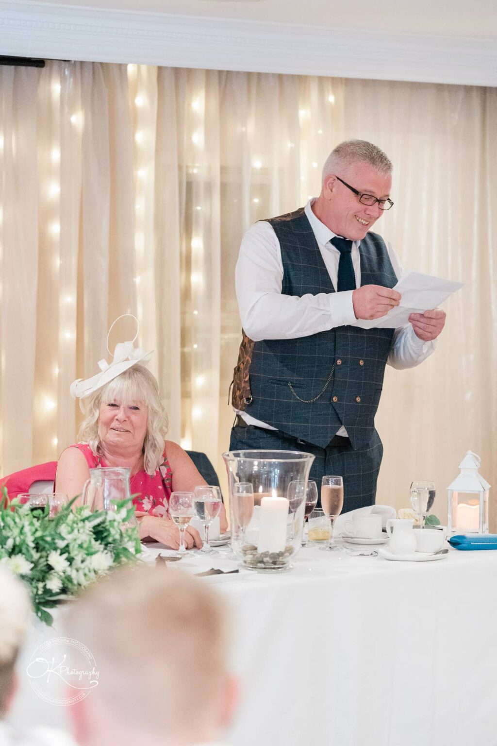 Santos Higham Farm Wedding Photography A man in a suit gives a speech at a wedding reception, while a woman in a pink dress and white hat sits next to him, smiling. The table is set with drinks, candles, and flowers, with a backdrop of light curtains and fairy lights.