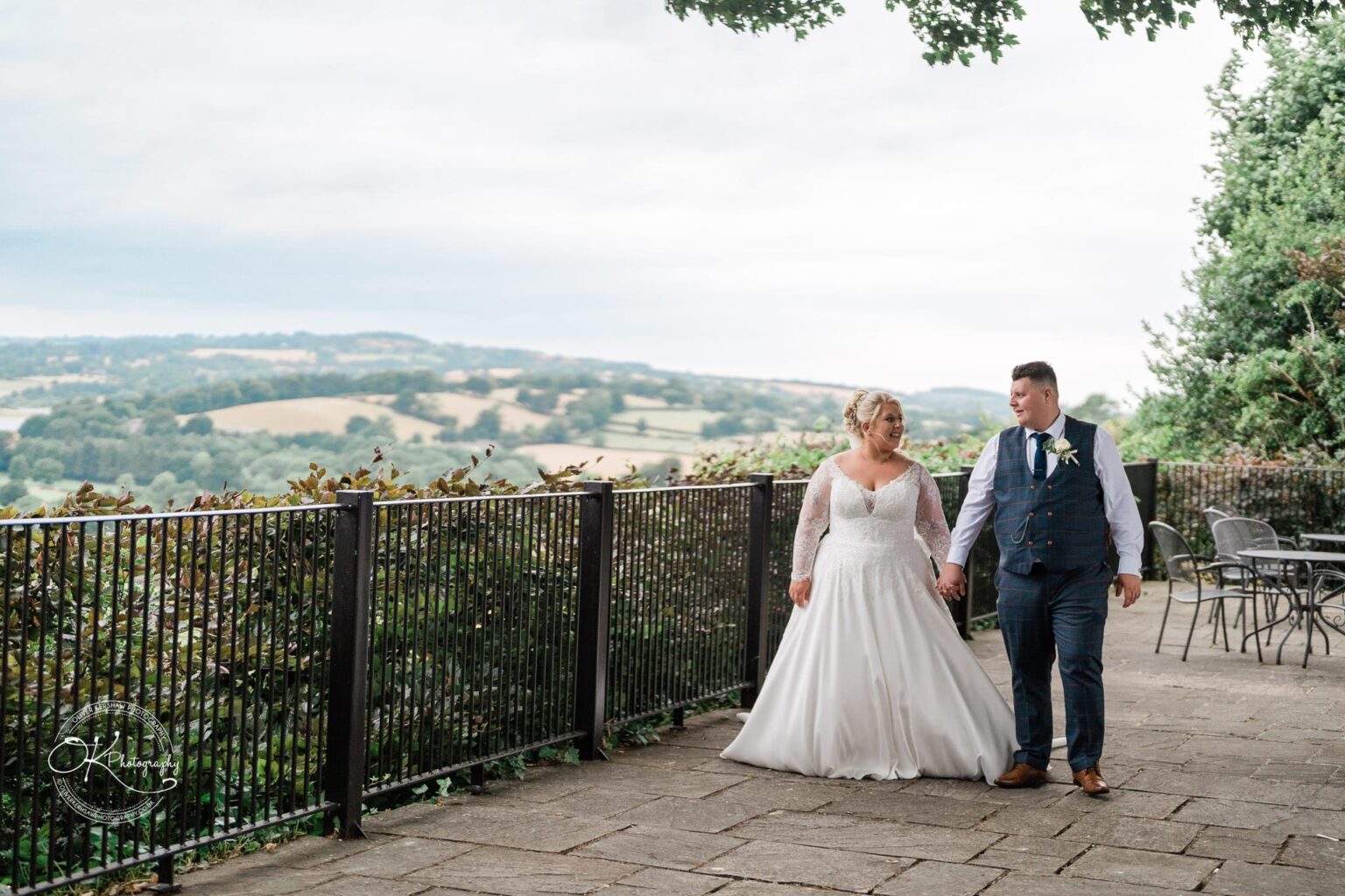 Santos Higham Farm Wedding Photography Bride and groom holding hands while walking on a patio with a scenic countryside view in the background.