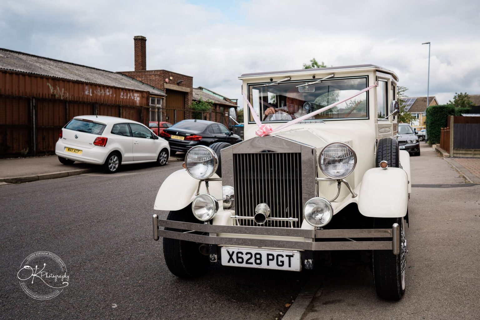 Shearsby Bath Wedding Photography A white vintage car with pink ribbon on its bonnet is parked on a street beside other modern cars.