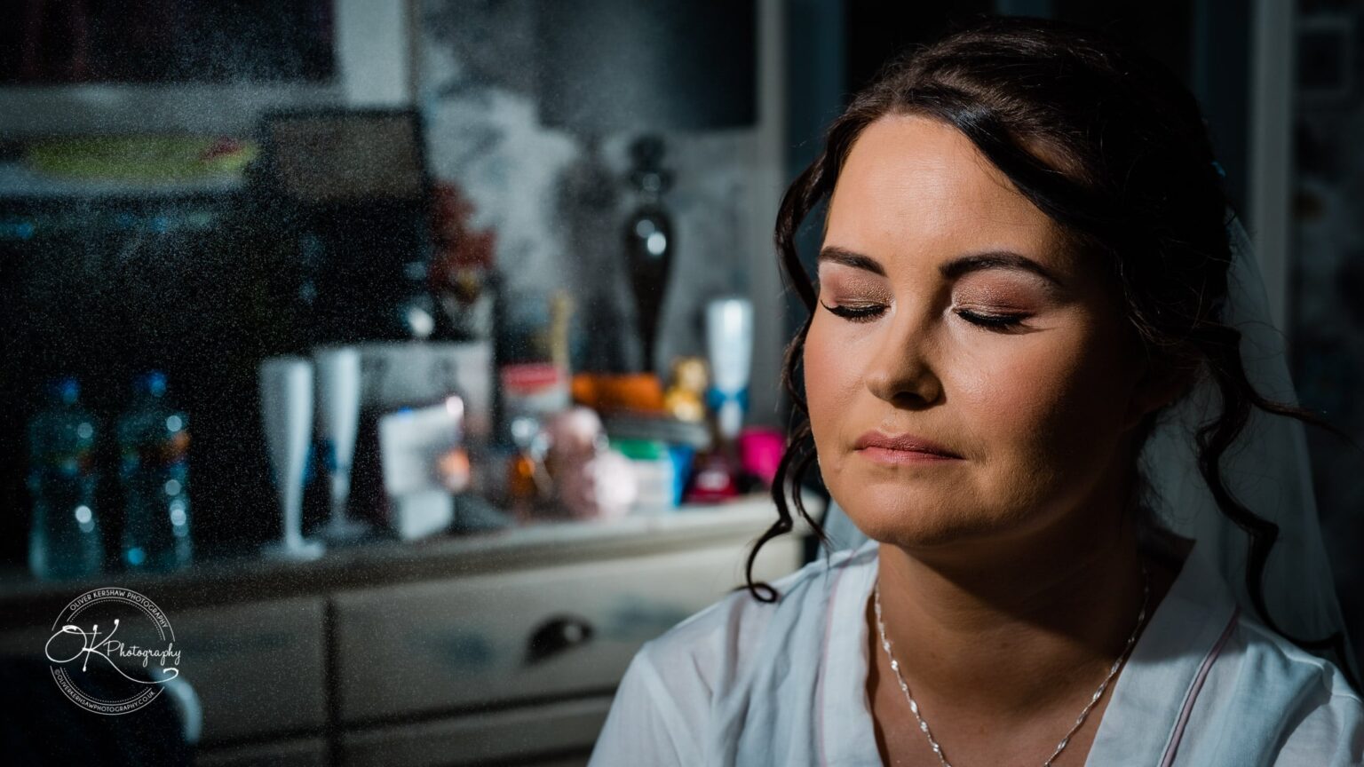 Shearsby Bath Wedding Photography A woman with closed eyes and styled hair, wearing a necklace, in front of a blurred background with various objects.