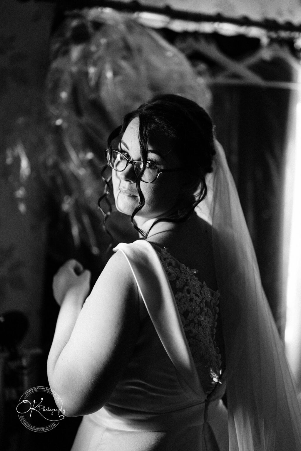 Shearsby Bath Wedding Photography Black and white portrait of a bride with glasses, gazing over her shoulder with a soft smile, wearing a dress with lace details and a flowing veil.