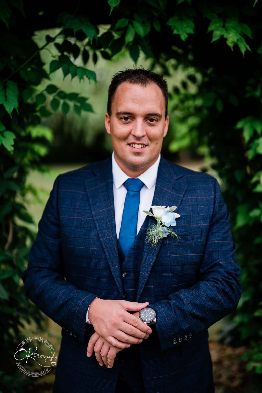 Shearsby Bath Wedding Photography A man in a blue checked suit with a blue tie and boutonnière, standing in front of green foliage.