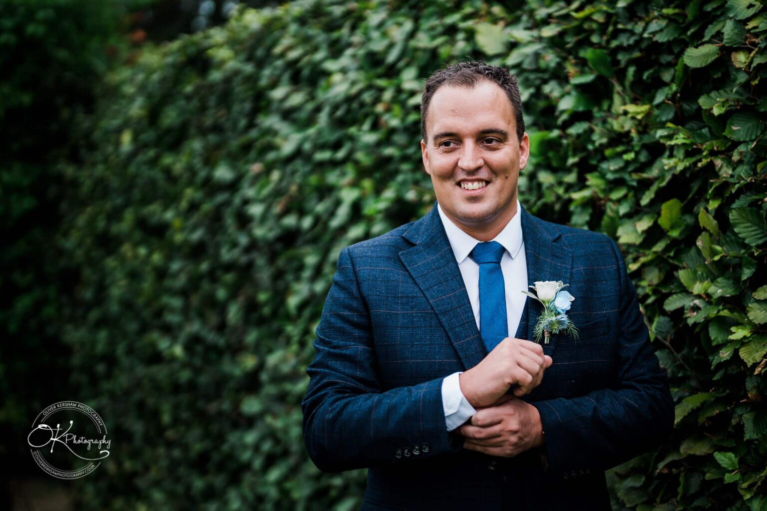 Shearsby Bath Wedding Photography Man in blue checkered suit and blue tie with a boutonniere, standing in front of a leafy hedge.