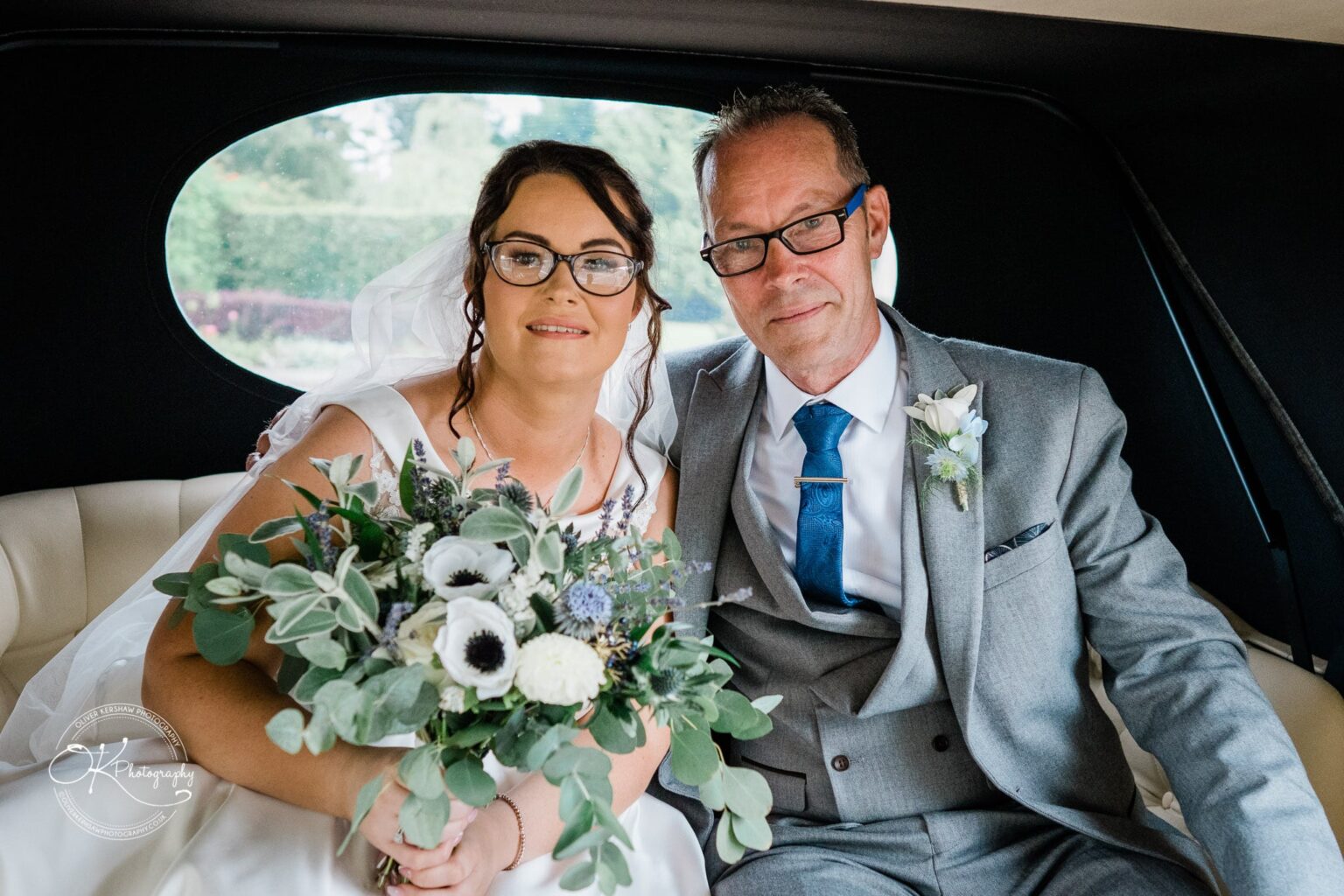 Shearsby Bath Wedding Photography A bride and an older man sitting inside a vehicle; she holds a bouquet of white and green flowers. Both are wearing glasses and smiling.