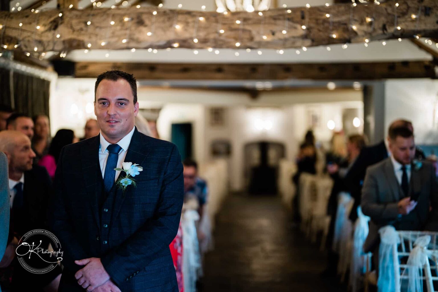 Shearsby Bath Wedding Photography A groom stands in an indoor wedding venue decorated with string lights and wood beams, surrounded by seated guests.