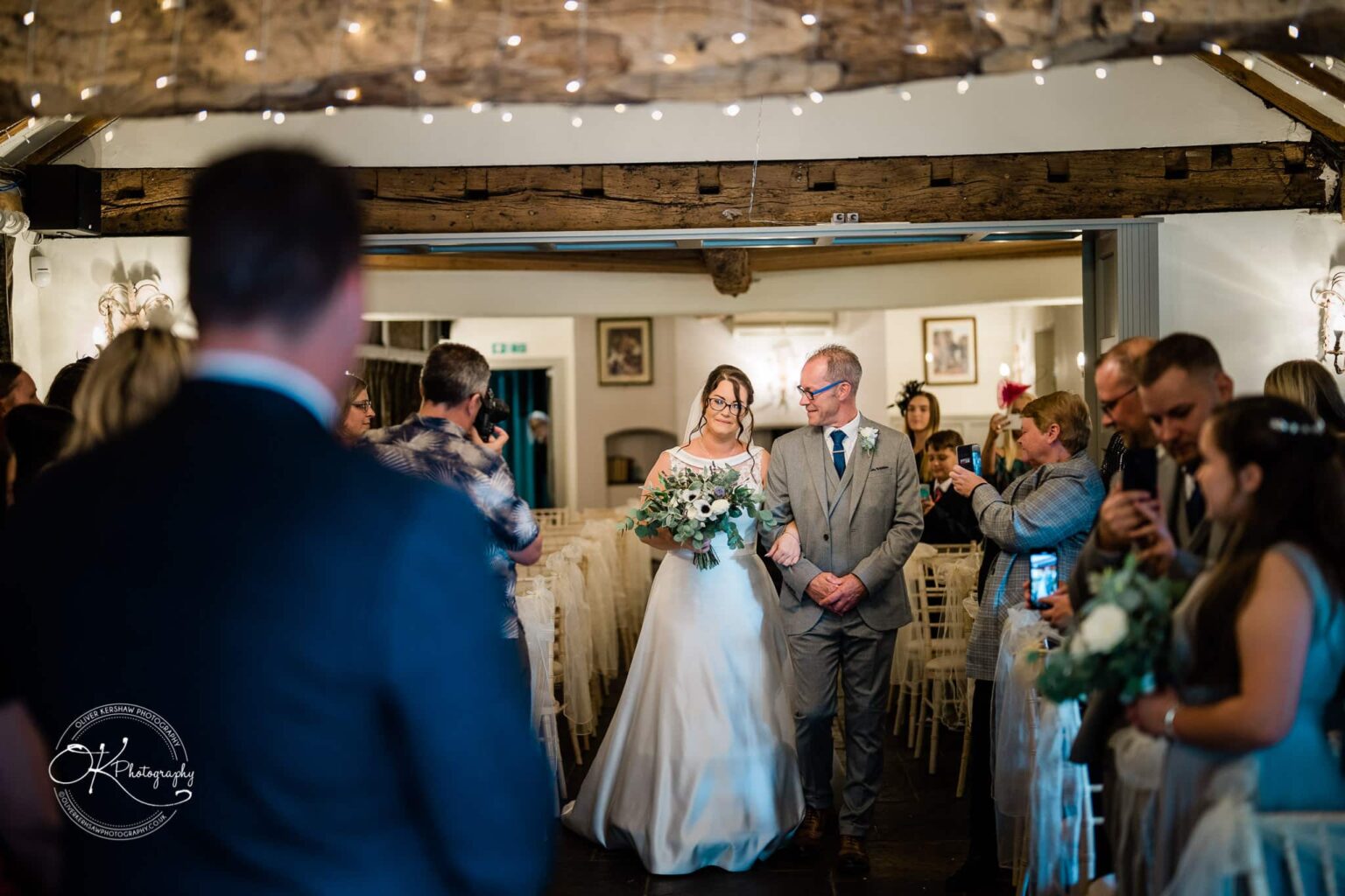 Shearsby Bath Wedding Photography A bride in a white gown holding a bouquet walks down the aisle with an older man in a grey suit, surrounded by guests taking pictures inside a rustic venue adorned with fairy lights.