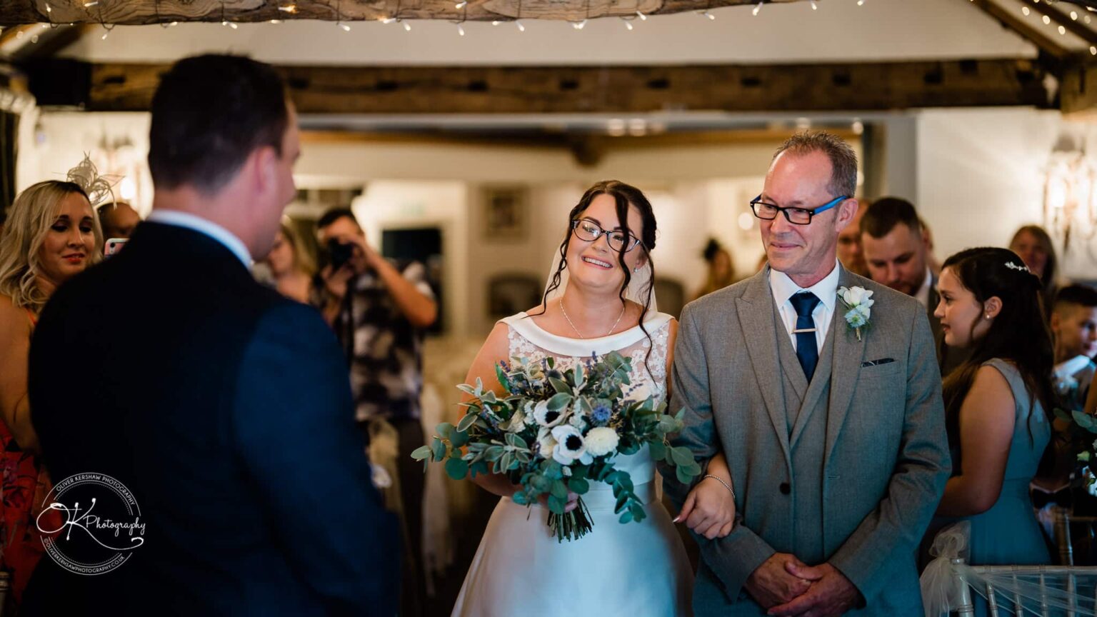 Shearsby Bath Wedding Photography A bride and her father smile as they walk down the aisle towards the groom, in a warmly lit indoor wedding venue decorated with fairy lights.