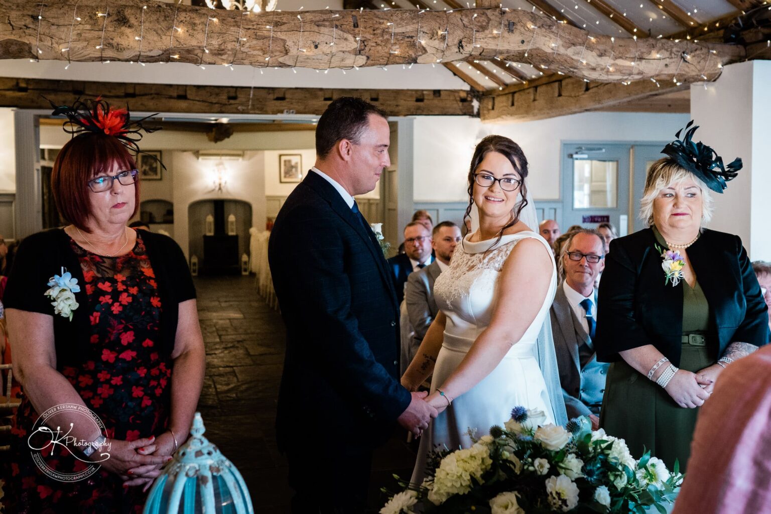 Shearsby Bath Wedding Photography A couple smiling and holding hands during a wedding ceremony, surrounded by guests seated in rows, inside a rustic venue with exposed wooden beams decorated with fairy lights.