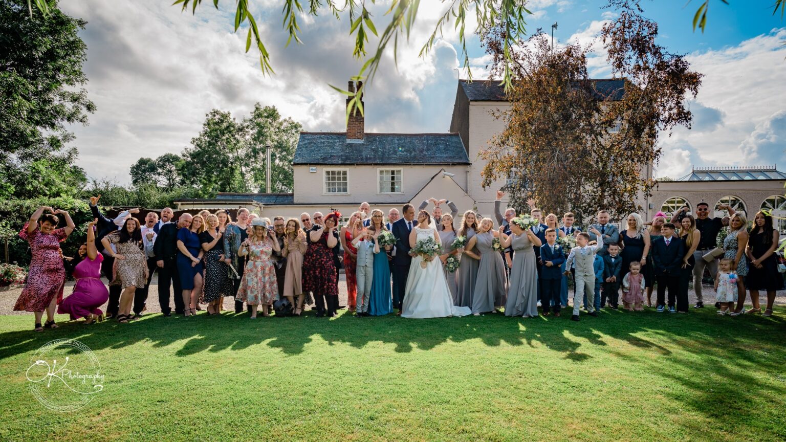 Shearsby Bath Wedding Photography A large wedding group posing for a photo in front of Shearsby Bath, a historical building with lush greenery and a bright sky in the background.