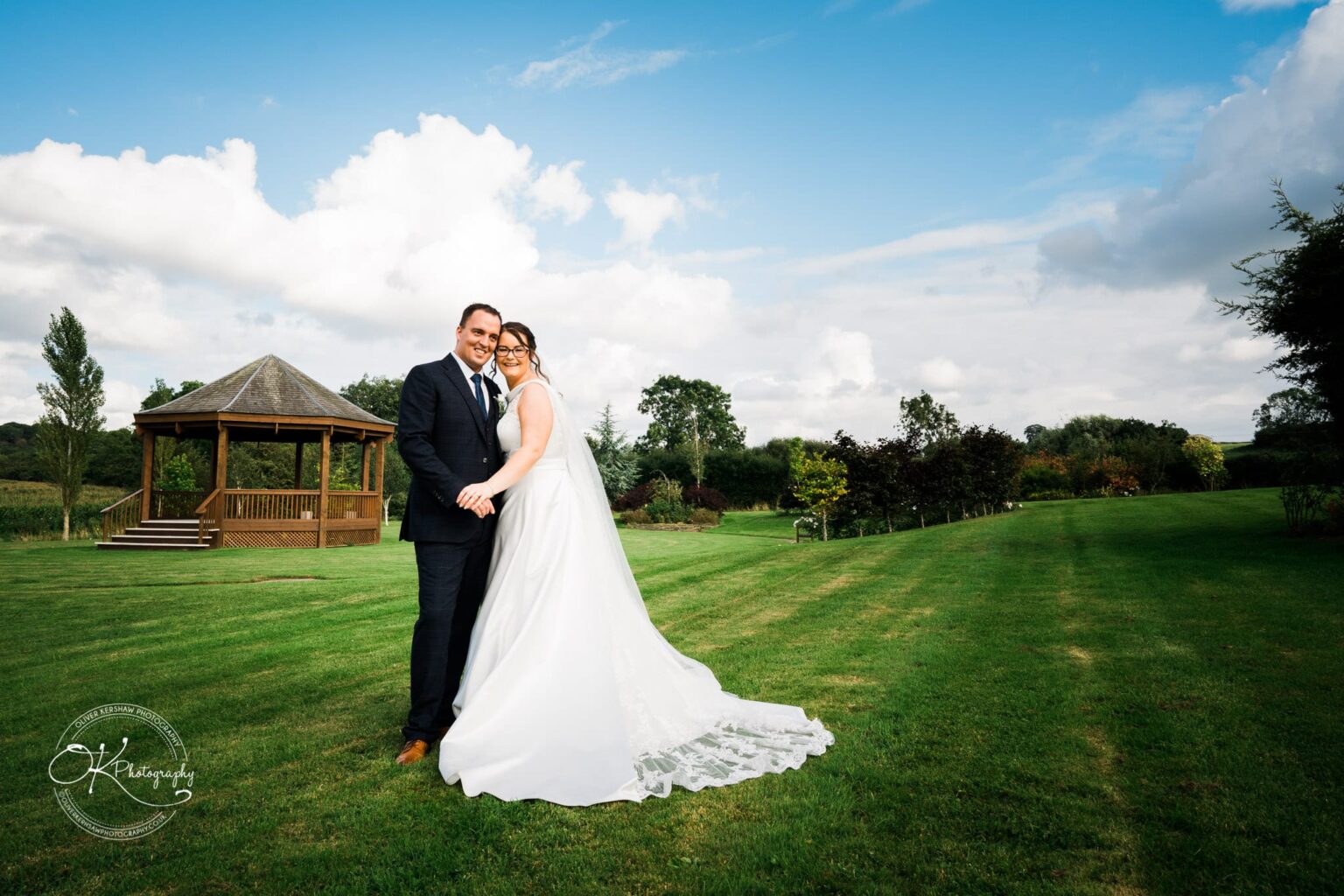 Shearsby Bath Wedding Photography A newlywed couple posing on a large green lawn with a wooden gazebo in the background and a partly cloudy sky.