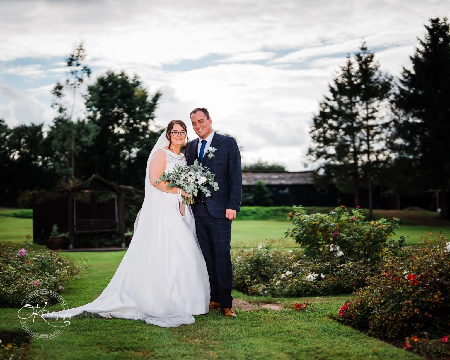 Shearsby Bath Wedding Photography Bride and groom in wedding attire posing outside with greenery and flowers in the background.