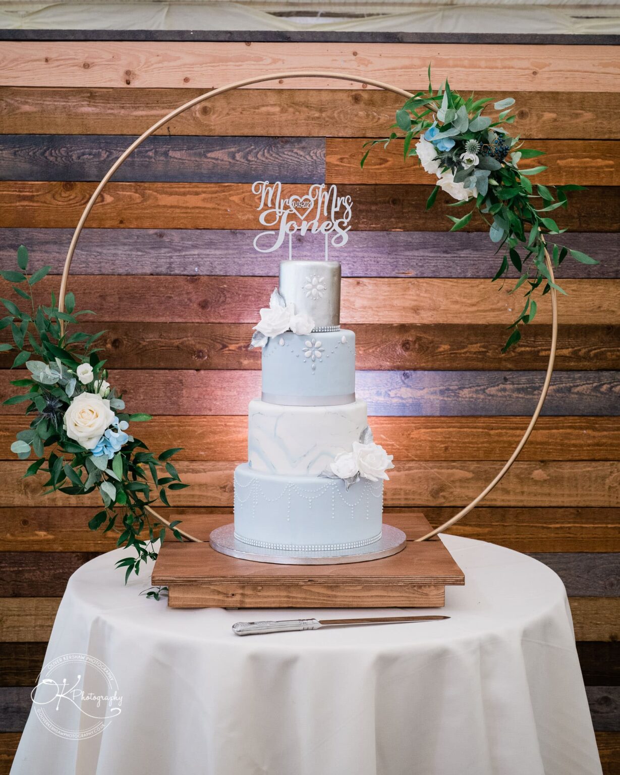Shearsby Bath Wedding Photography A three-tiered blue and white wedding cake with floral decorations, displayed on a round table with a wooden background and floral arch.