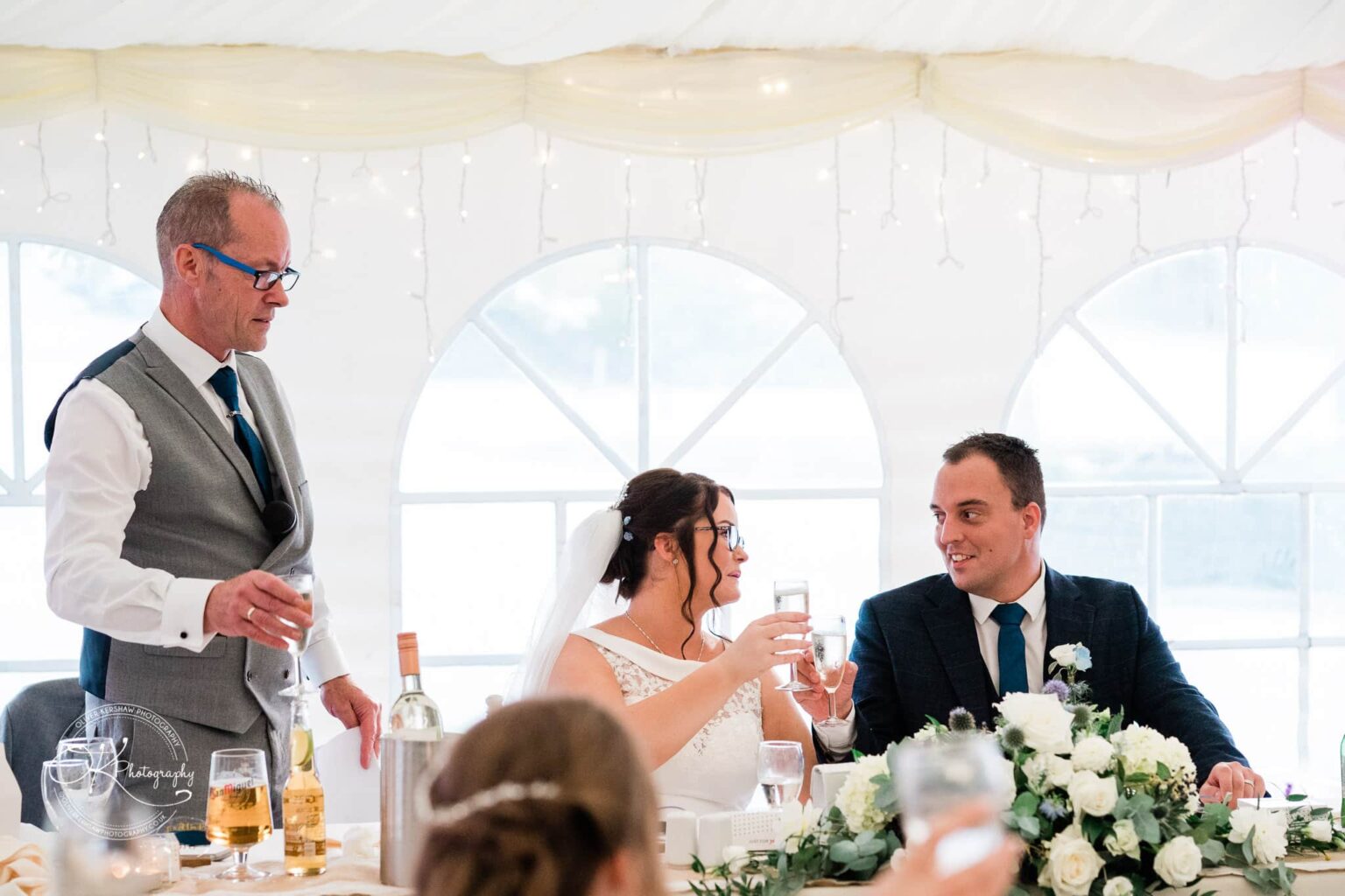Shearsby Bath Wedding Photography A wedding couple toasting with a seated man in a grey vest and blue tie standing next to them, with white floral decorations and a bottle of wine on the table in a white tent.