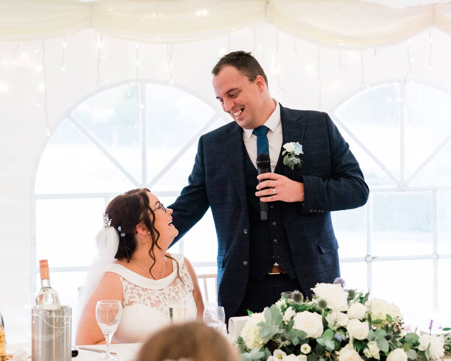 Shearsby Bath Wedding Photography A couple at their wedding reception, with the groom giving a speech while the bride looks up and smiles at him.