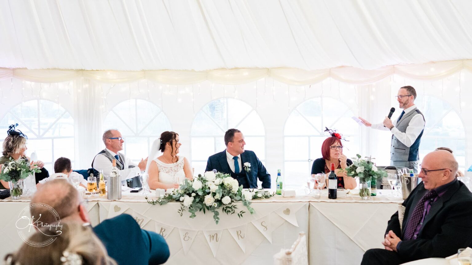 Shearsby Bath Wedding Photography Wedding reception at Shearsby Bath with guests seated at a long table, decorated with flowers and a 'Mr & Mrs' banner. One man is standing, giving a speech with a microphone.