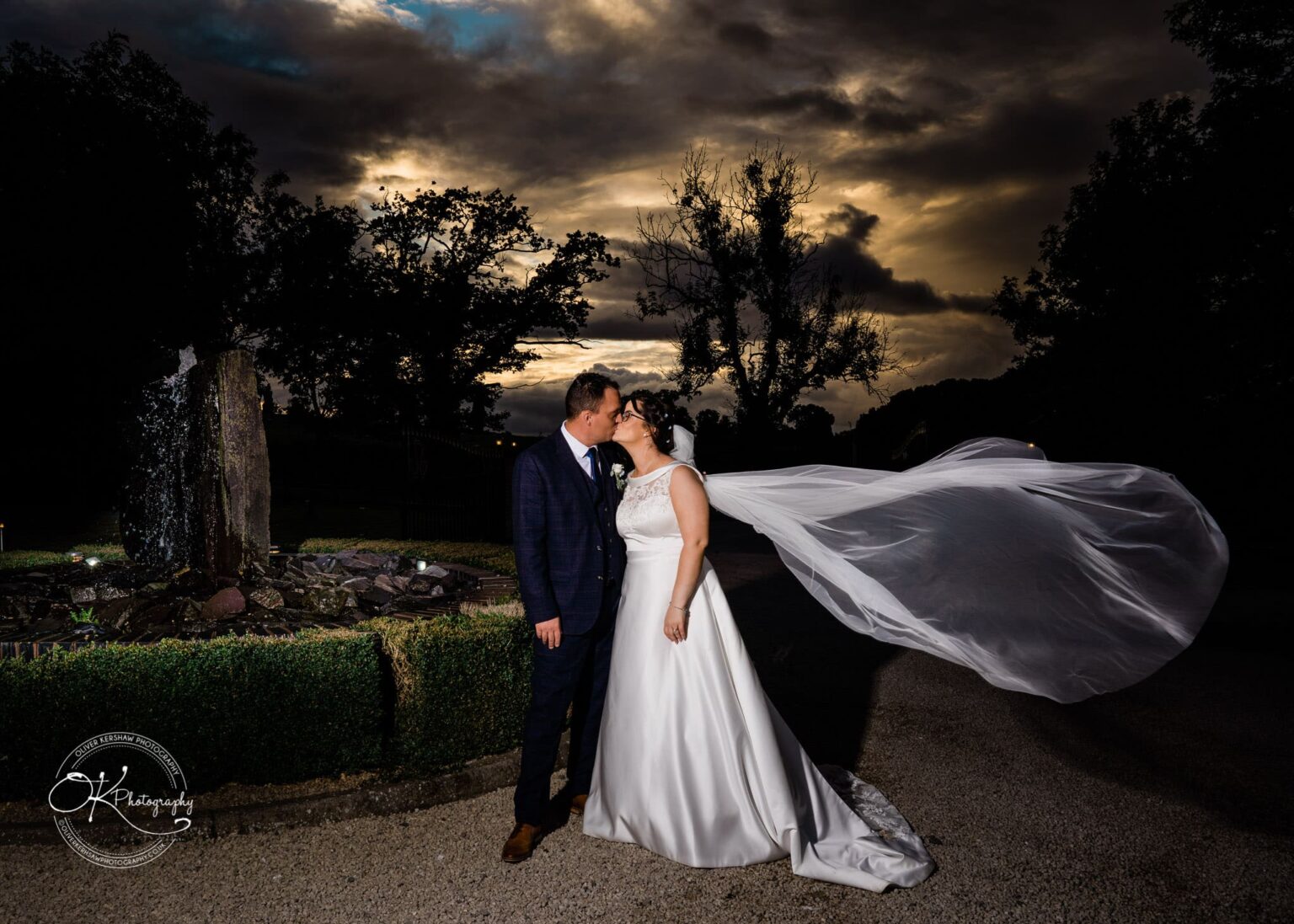 Shearsby Bath Wedding Photography A bride and groom share a kiss outside, with the bride's veil flowing in the wind against a dramatic cloudy sky backdrop.