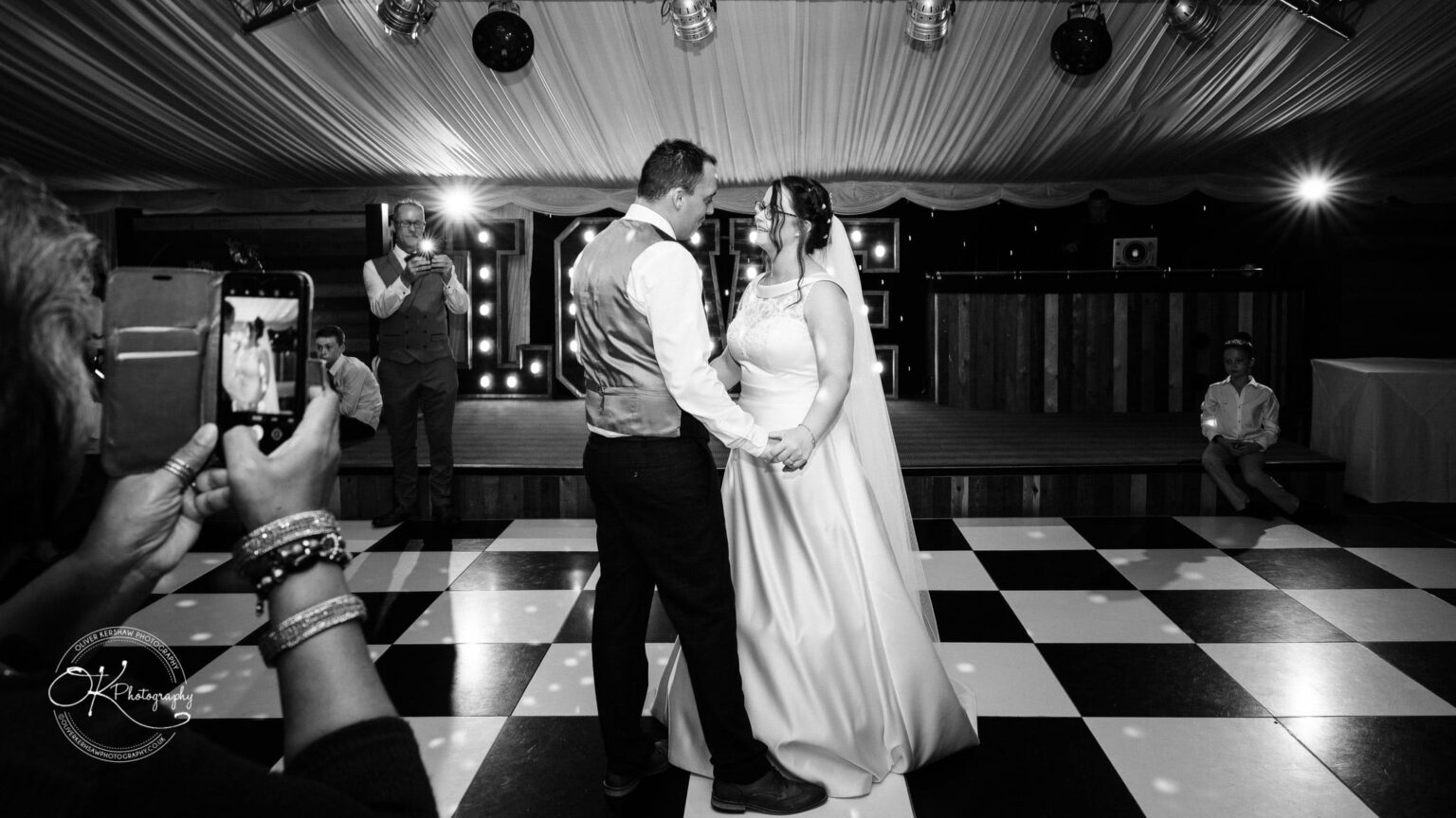 Shearsby Bath Wedding Photography A bride and groom dance while guests take photos at a wedding reception on a black and white checkered dance floor under a tent with lights.
