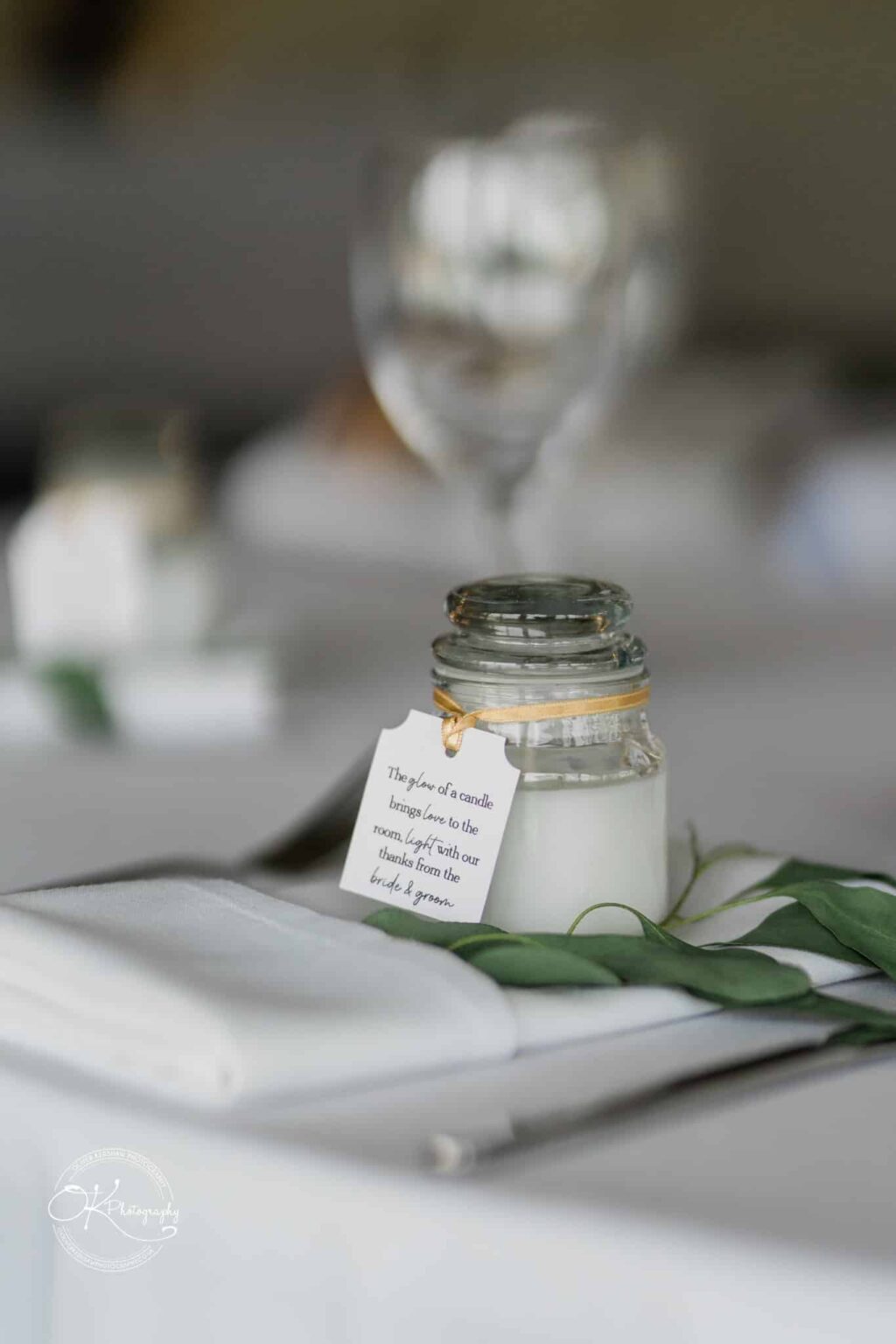 A close-up of a small glass jar candle with a tag, placed on a table with a white napkin and green foliage, and blurred glassware in the background.