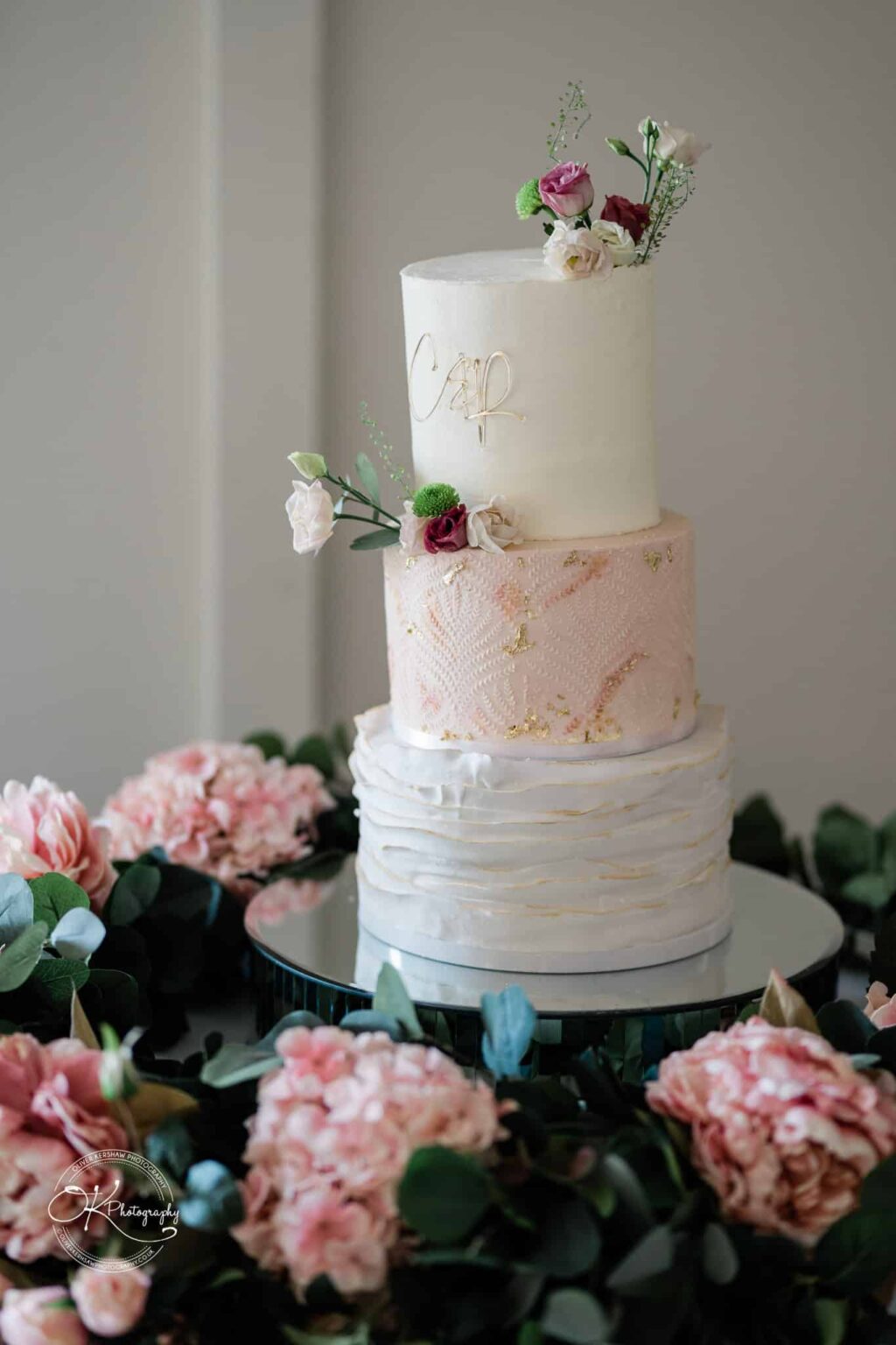 A three-tier wedding cake decorated with light pink and white frosting and adorned with flowers, placed on a reflective surface with pink flowers and greenery around the base.