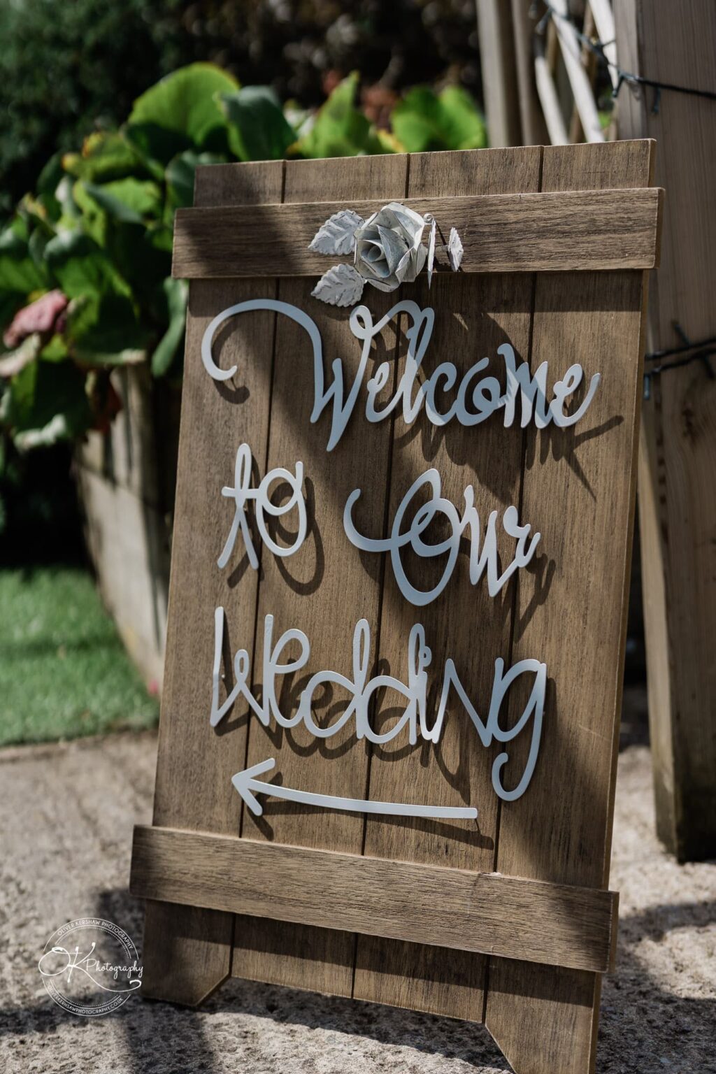 Wooden wedding sign with the words "Welcome to Our Wedding" and an arrow pointing left, decorated with a white flower and leaves.