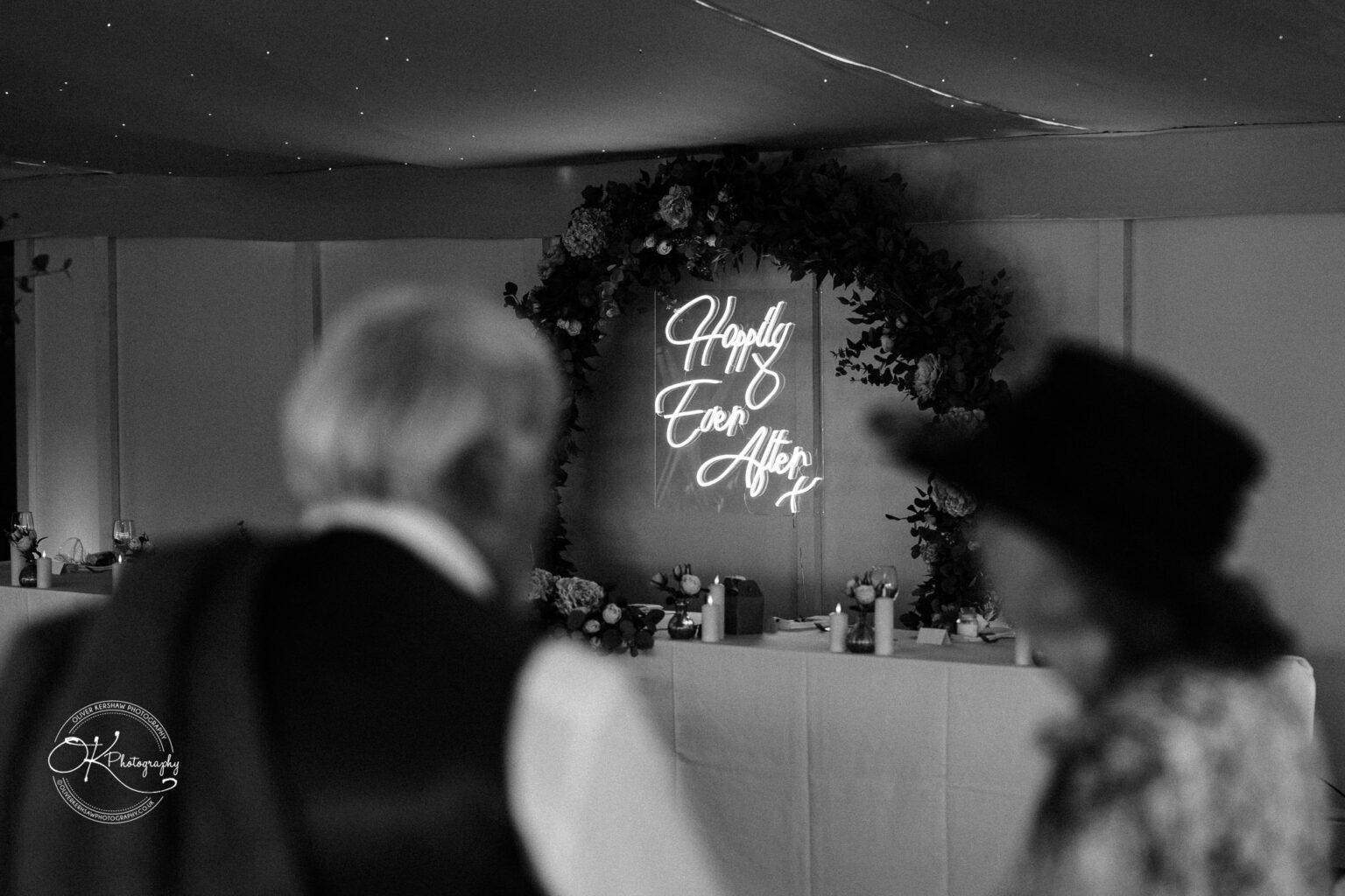 Black and white photo of an indoor wedding venue with a neon sign reading "Happily Ever After" surrounded by floral decorations, and two out of focus figures in the foreground.