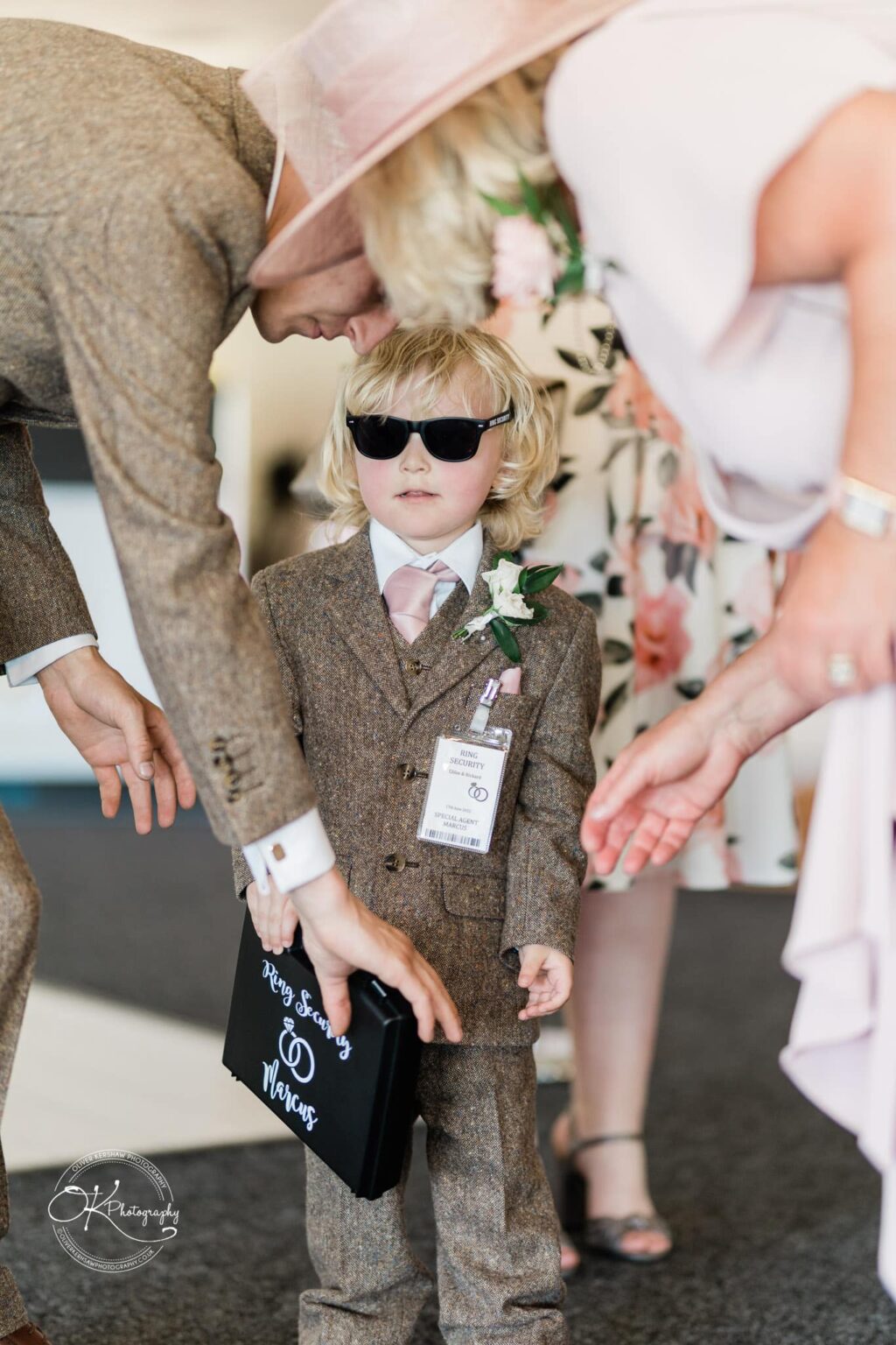 A young boy in a brown tweed suit, wearing sunglasses and holding a briefcase labeled "Ring Security Marcus," stands between two adults bending over him.