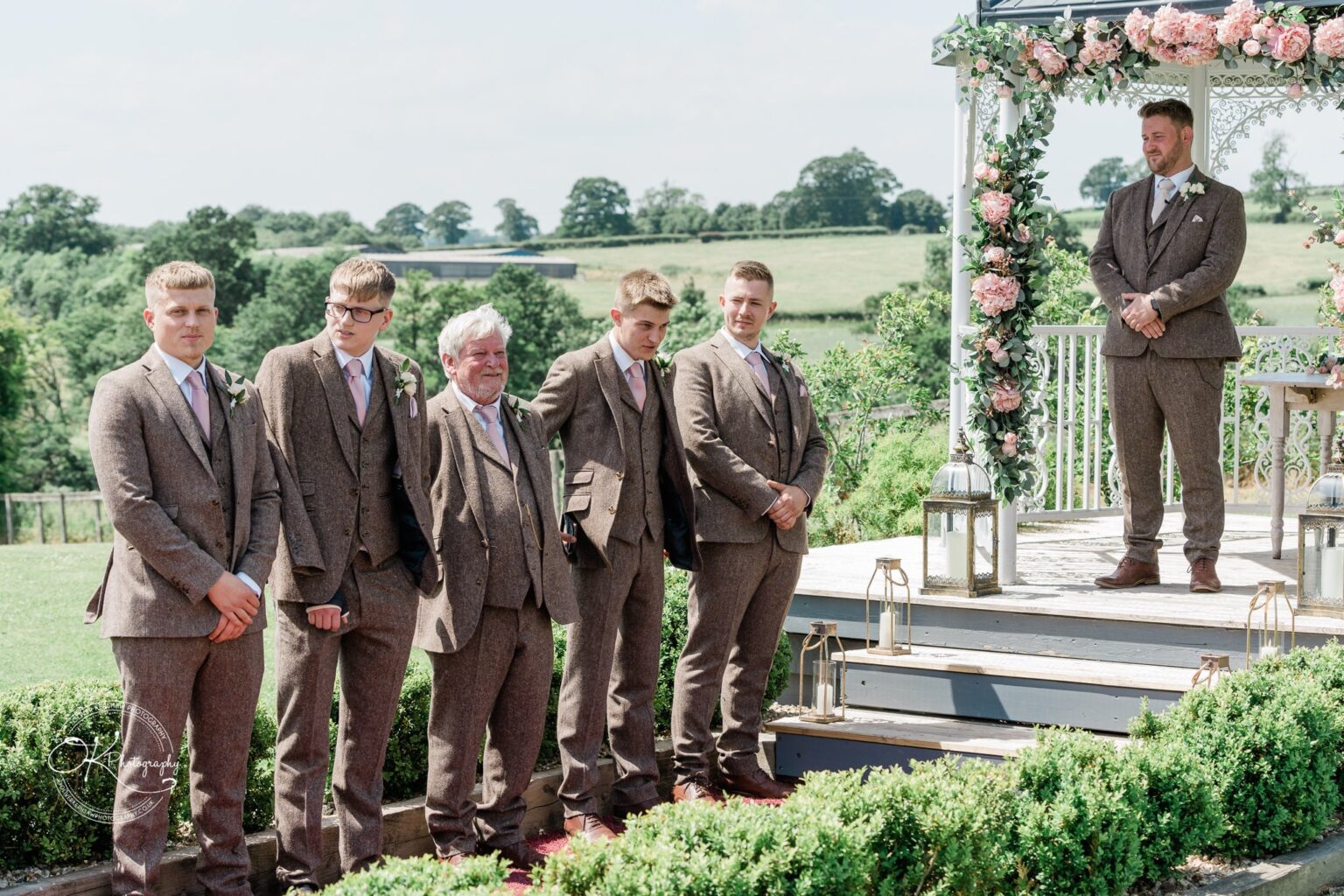 A group of men in brown suits standing in front of a decorated outdoor wedding gazebo at Shottle Hall, surrounded by green fields and trees.