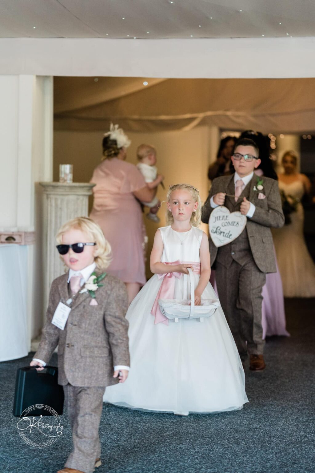 Young girl in a white dress and young boys in suits walking down an aisle at a wedding, one boy holding a briefcase and another holding a heart-shaped sign.