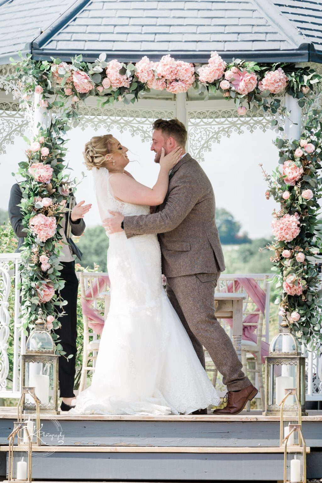 Bride and groom sharing a kiss at an outdoor wedding ceremony under a gazebo decorated with pink flowers and greenery.