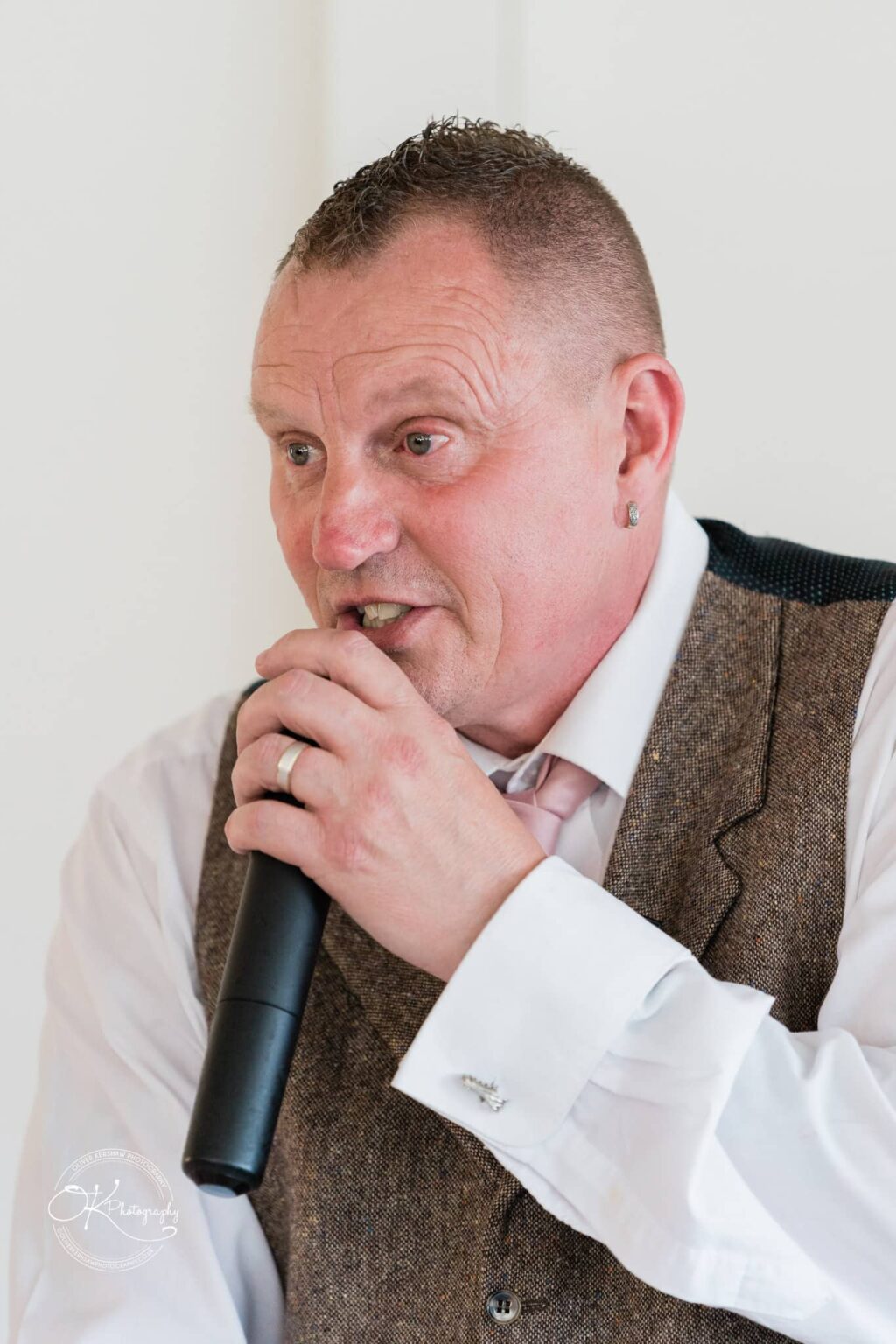 A man in a white shirt, tie, and brown vest speaks into a microphone.