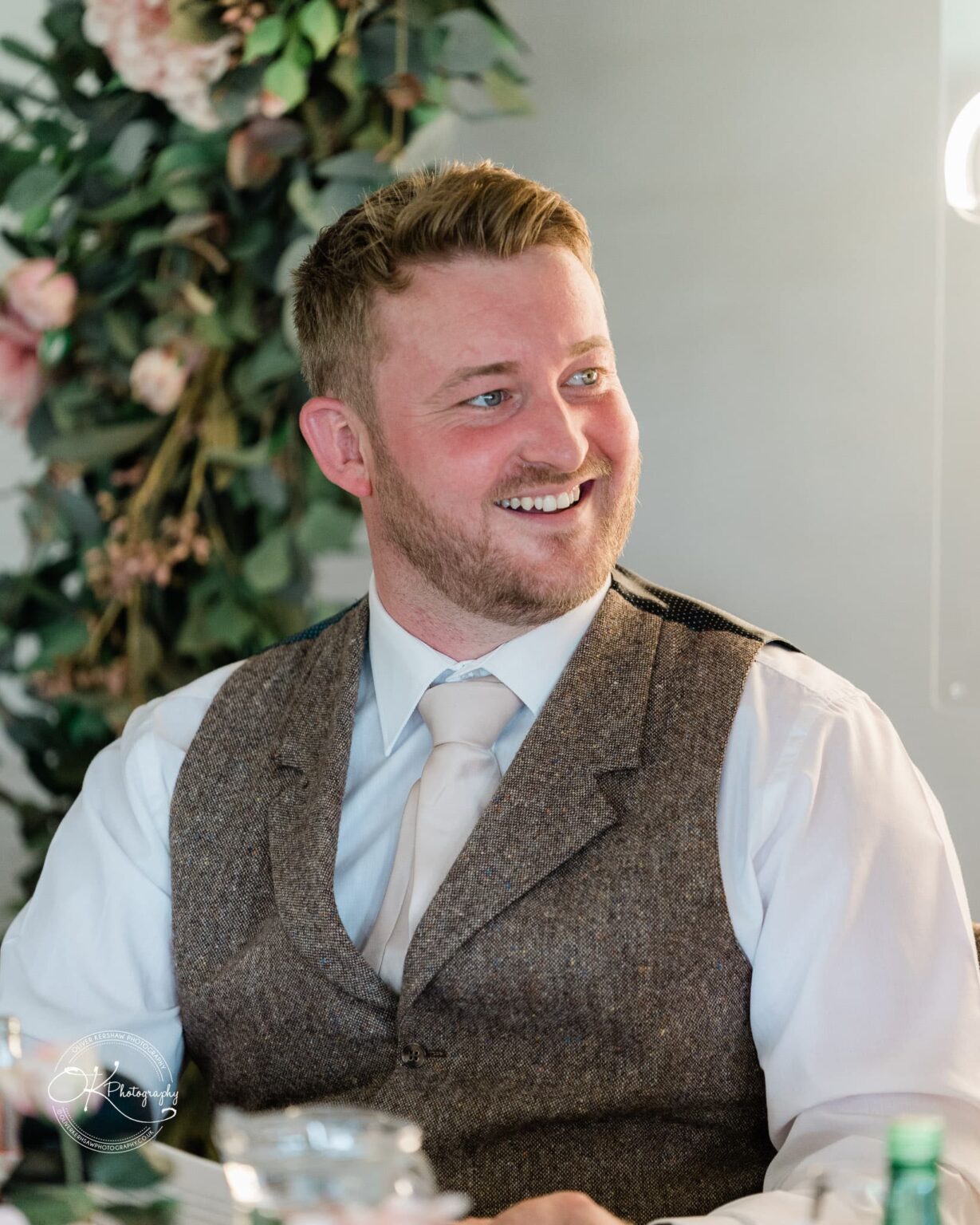 A man with short hair wearing a white shirt, tie, and brown vest, smiling and sitting indoors with greenery in the background.