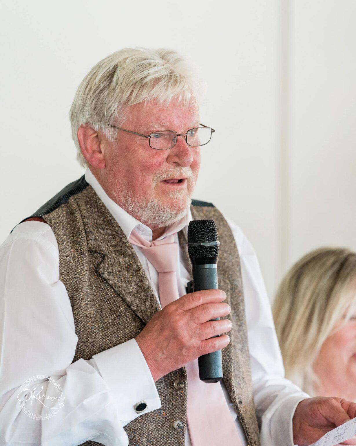 An elderly man with white hair and glasses speaks into a microphone, wearing a vest, white shirt, and pink tie.