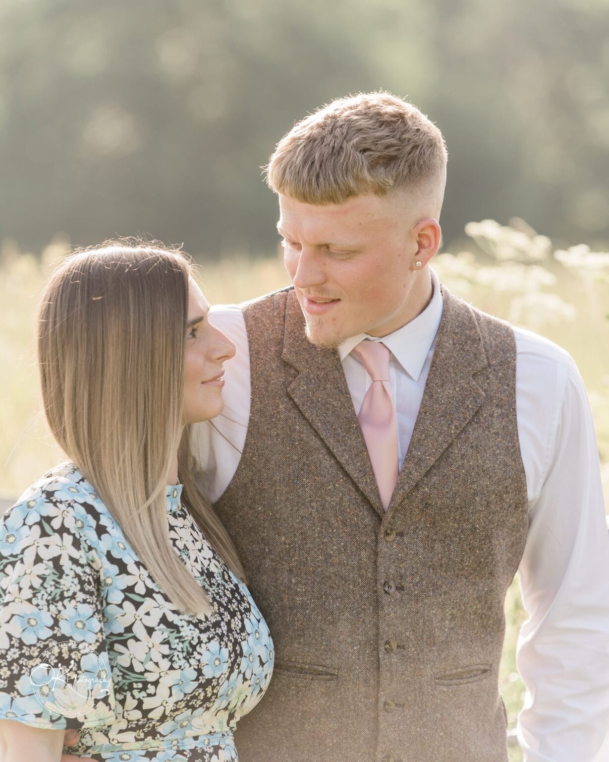 A couple standing close together, looking at each other lovingly; the man is wearing a tweed vest, white shirt, and pink tie, while the woman is in a floral dress. The background is a blurred, sunlit outdoor scene.