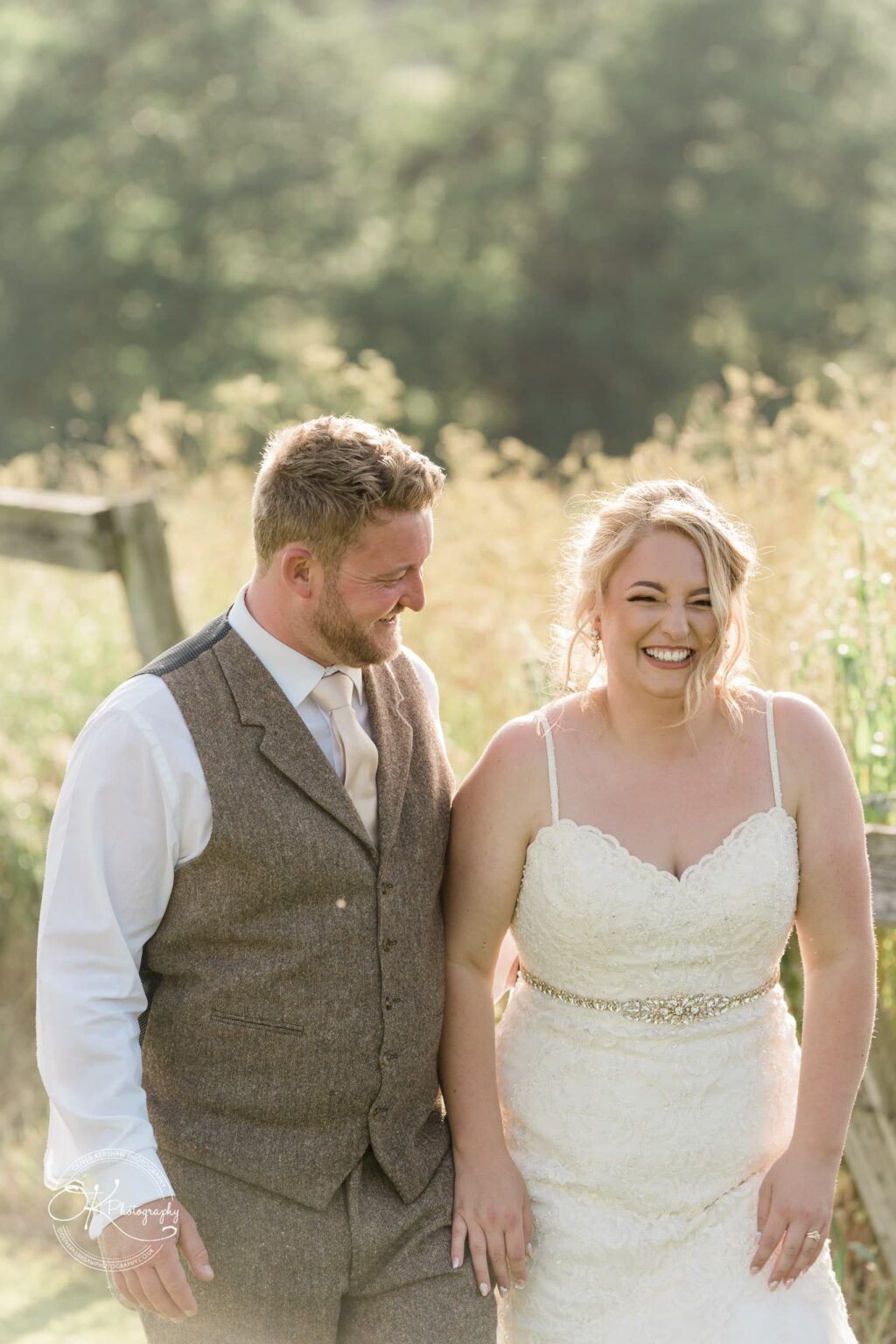 A bride and groom smiling and holding hands outdoors, with greenery in the background.