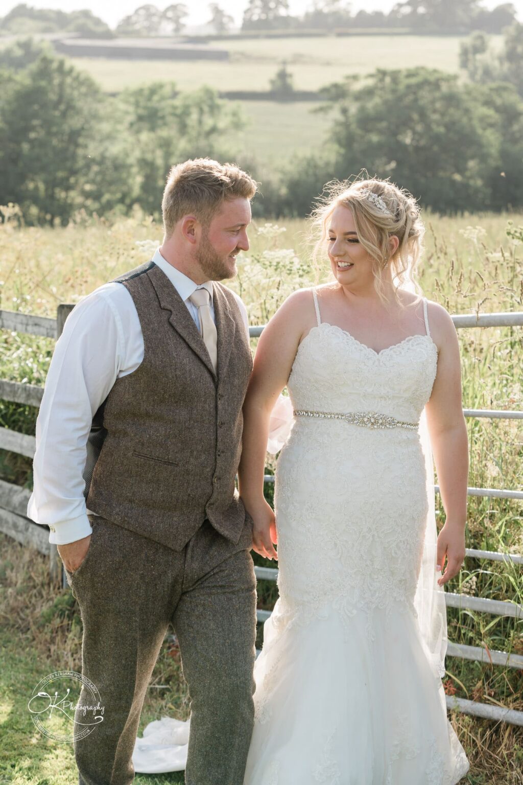 A bride and groom holding hands and walking outdoors in a grassy field, with trees and hills in the background.