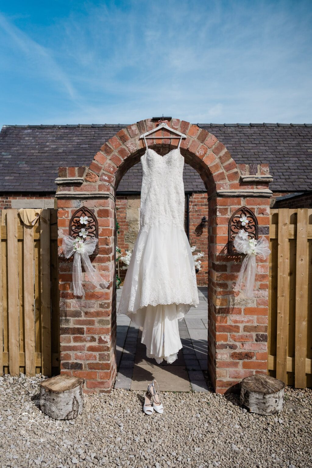 A white lace wedding dress hanging on a brick archway with white decorative ribbons, and a pair of white heels on the ground beneath it.