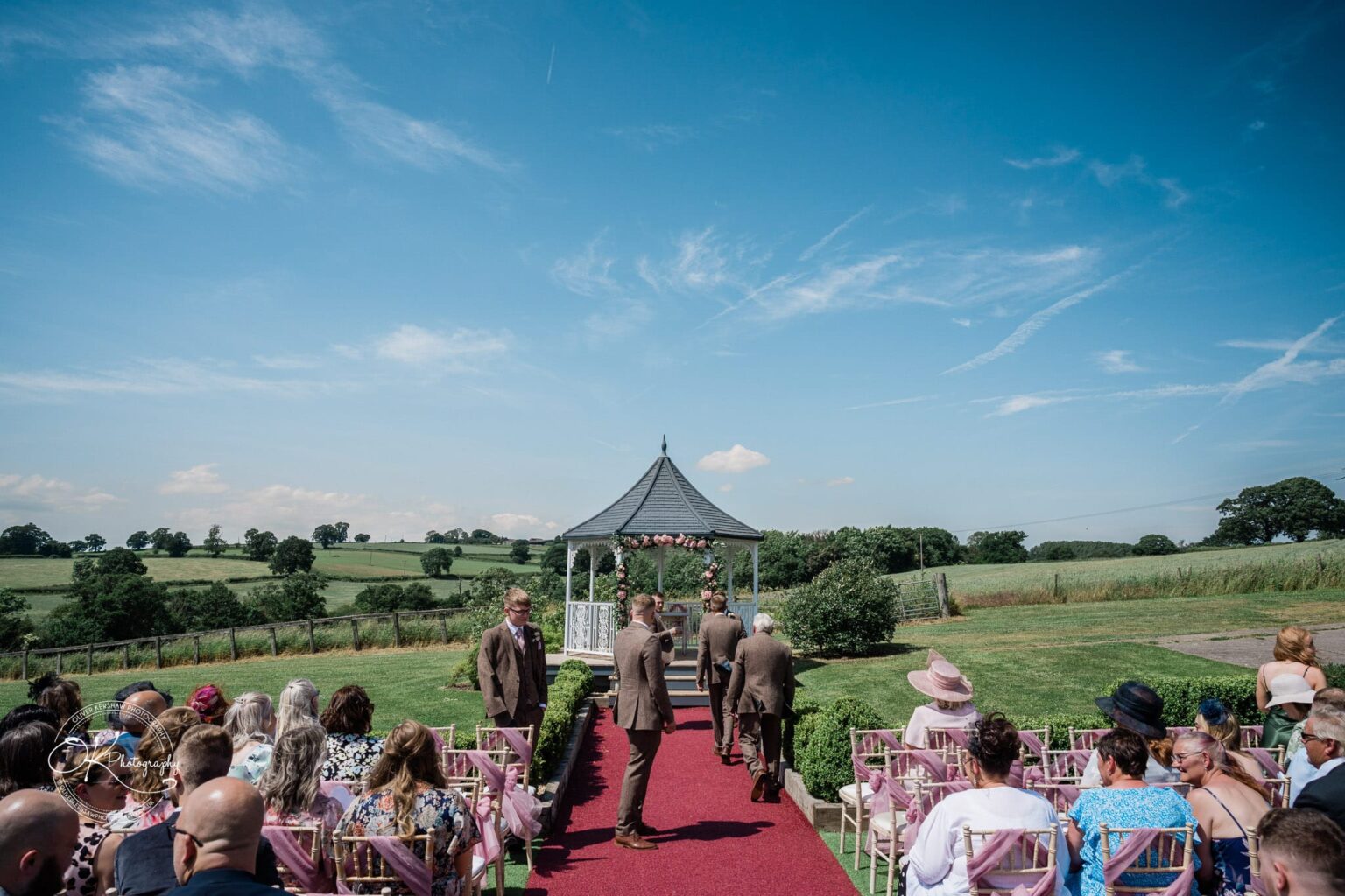 Outdoor wedding ceremony at Shottle Hall with guests seated and groomsmen walking towards an ornate gazebo, set against a backdrop of rolling green fields and a clear blue sky.