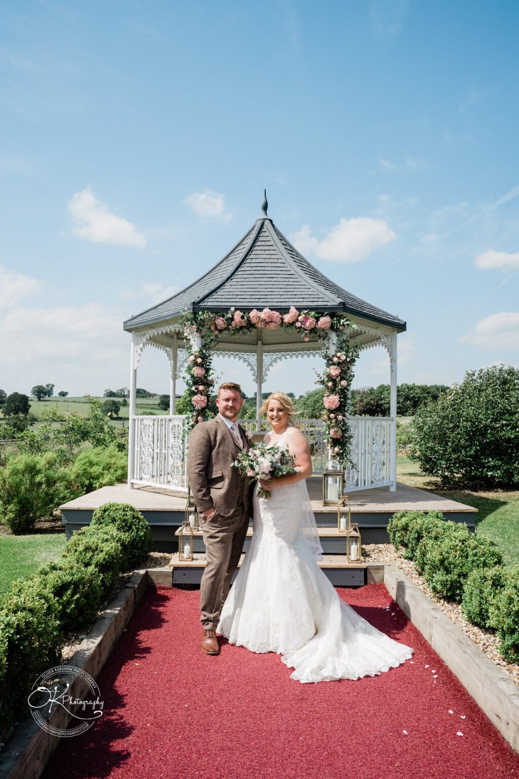A bride and groom stand in front of a decorated gazebo at Shottle Hall, with a scenic countryside backdrop on a sunny day.