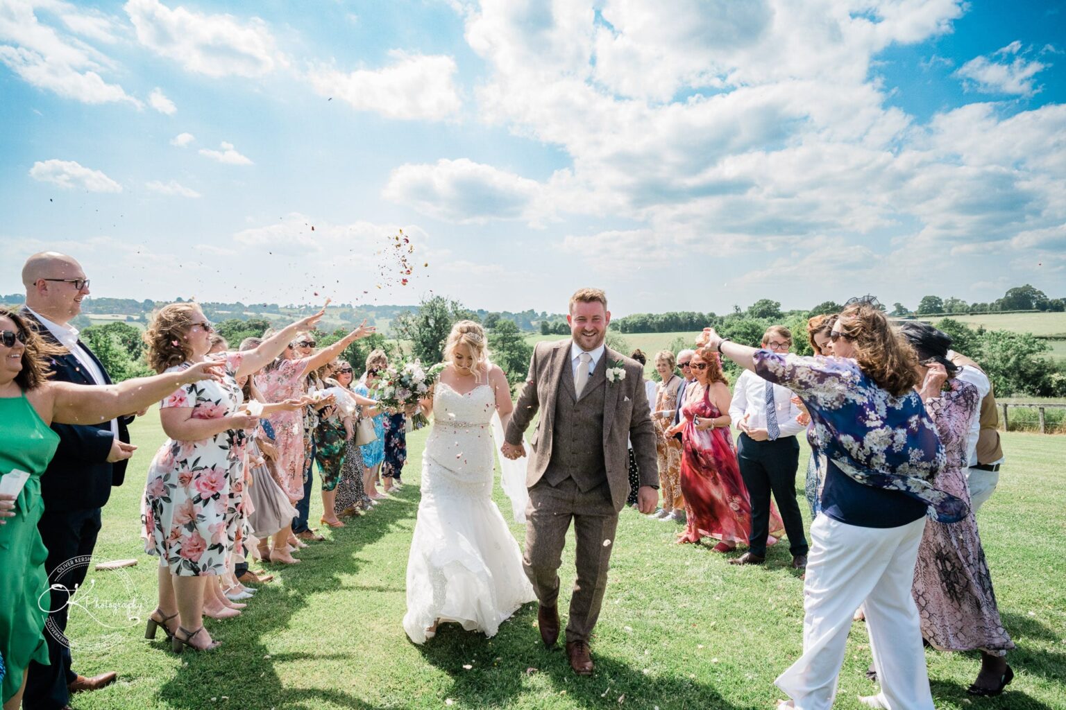 A wedding couple walking on a grassy lawn with guests throwing confetti, under a partly cloudy blue sky in a picturesque rural location.