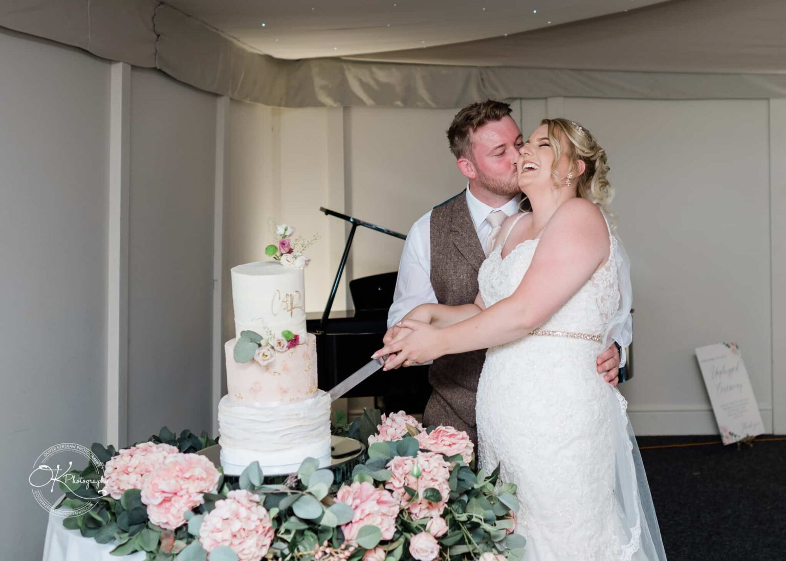 A newlywed couple smiling as they cut their wedding cake, which is adorned with flowers and on a table surrounded by pink flowers and greenery.