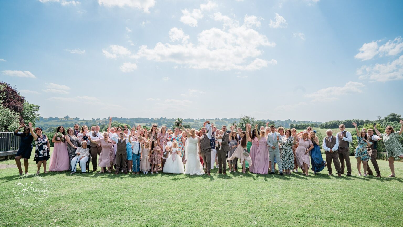 A large wedding group stands on a lawn with a scenic countryside background, posing and cheering under a bright blue sky at Shottle Hall.