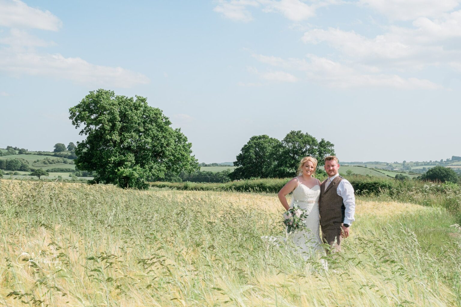 A couple standing in a grassy field with trees and rolling hills in the background under a partly cloudy sky.