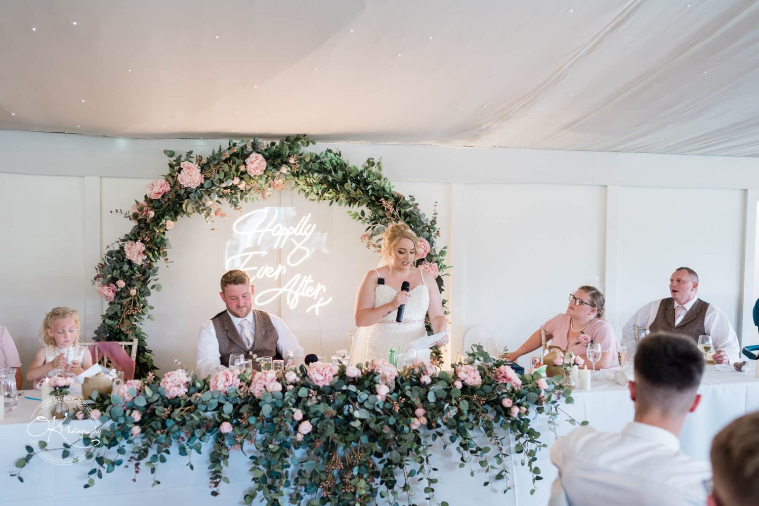 Bride and groom seated at a wedding table decorated with a floral arch and a neon sign that reads "Happily Ever After," with guests nearby.