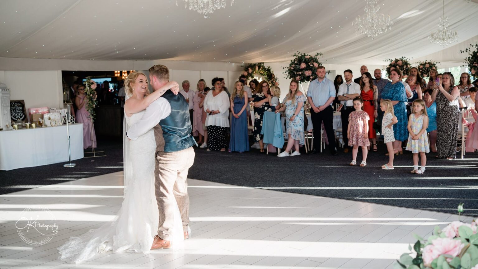 Newlywed couple dancing in a decorated marquee, surrounded by seated and standing guests watching them.