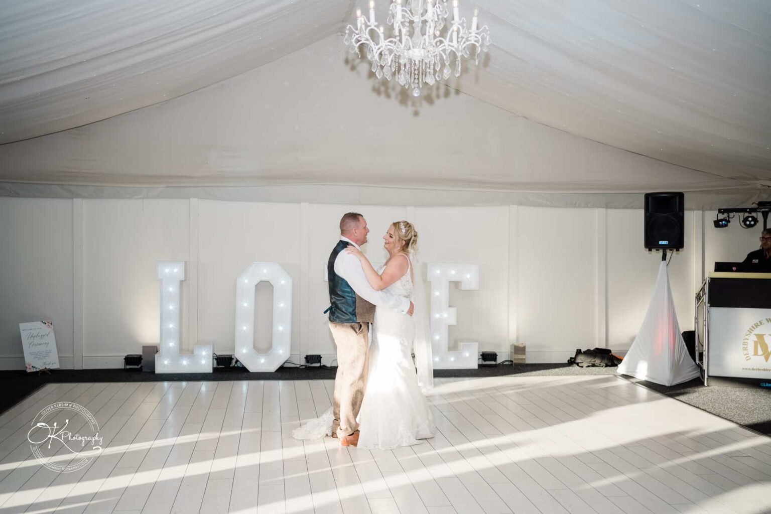 A bride and groom dancing on a white wooden floor beneath a chandelier, with illuminated "LOVE" letters in the background.