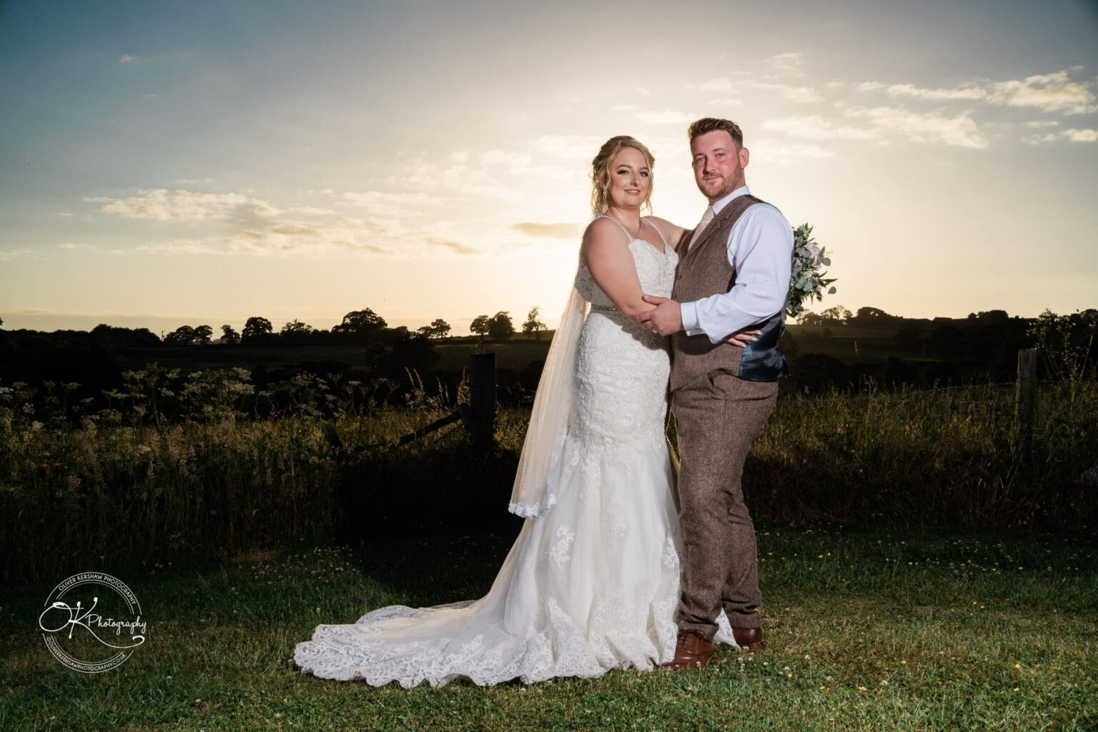 A bride and groom in wedding attire pose outdoors with a scenic countryside backdrop during sunset.