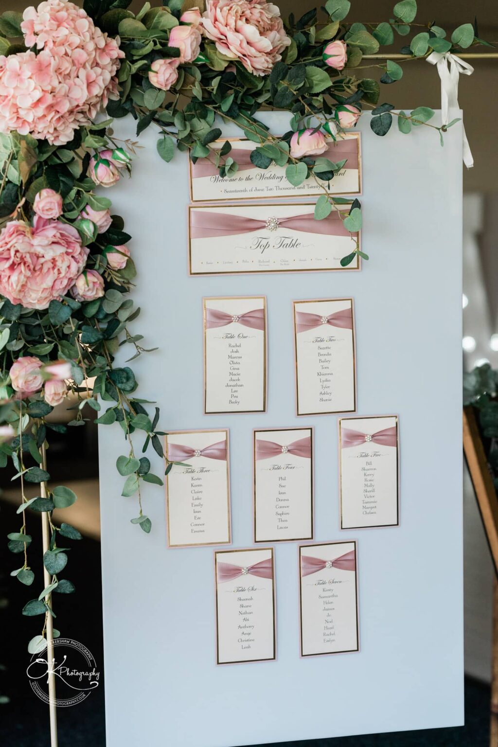 A wedding seating chart decorated with pink roses and greenery.