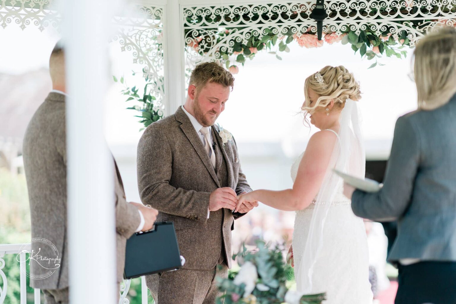 A bride and groom exchanging rings during an outdoor wedding ceremony under a decorated gazebo.