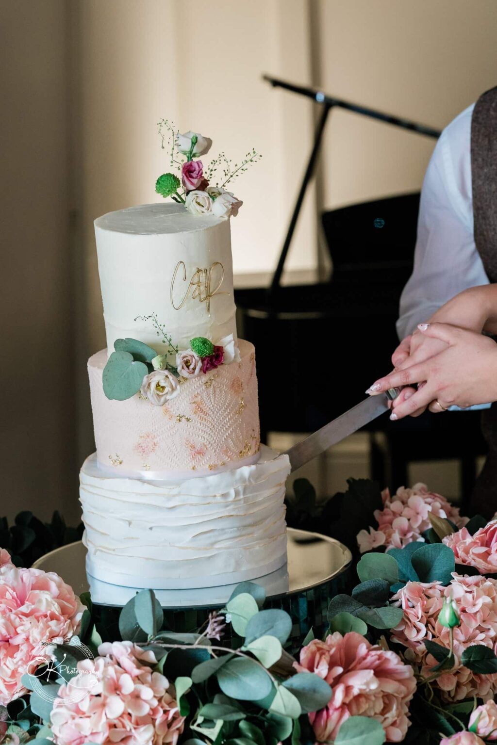 A three-tiered wedding cake decorated with flowers and golden accents being cut by a couple, surrounded by pink and white flowers.